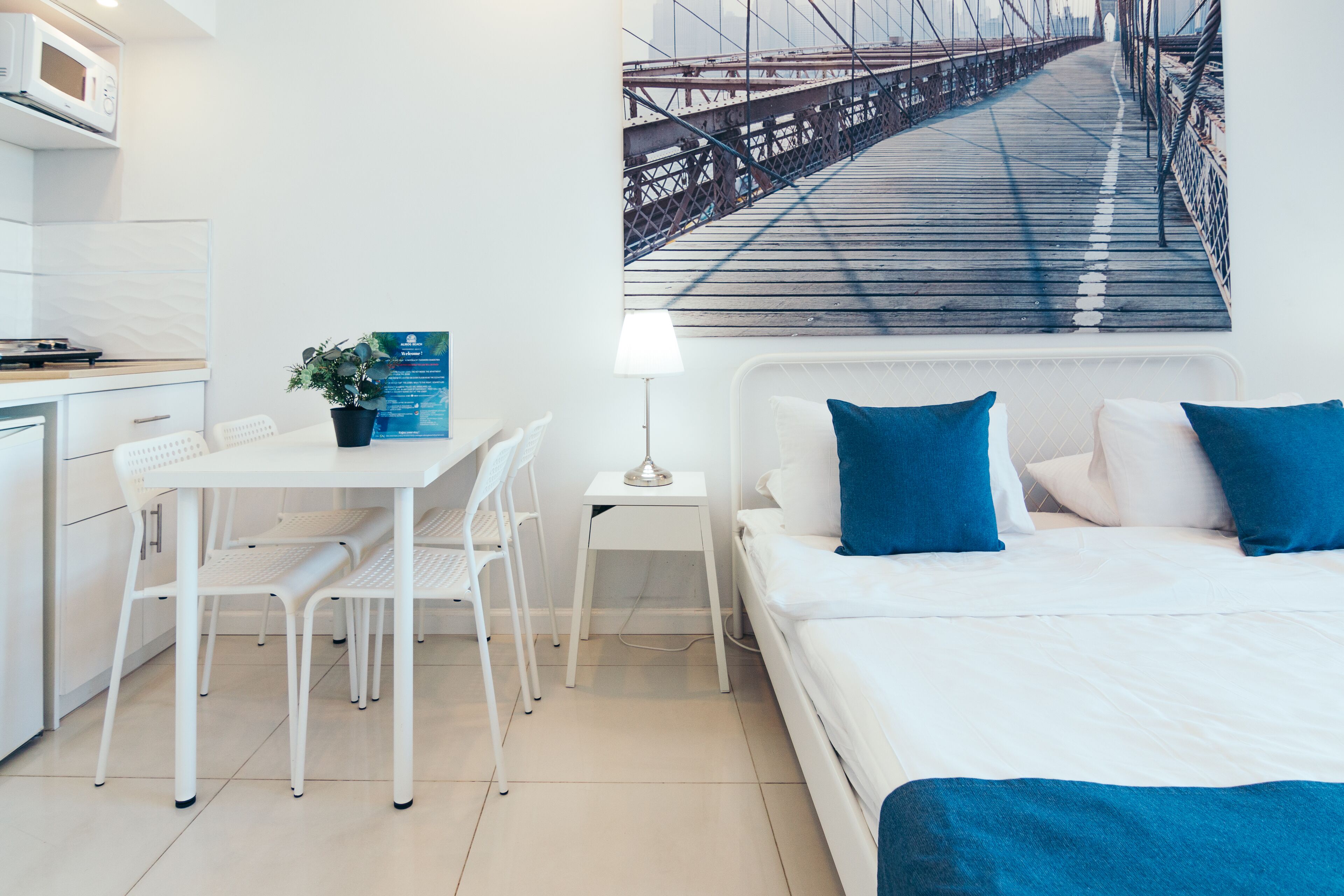 A modern hotel room with a bed, a desk, a chair, a small table, a lamp, and a large window with a cityscape view.