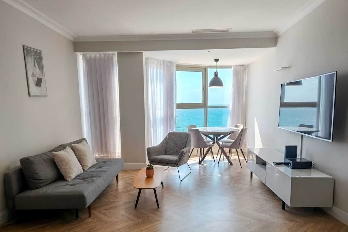 A modern living room with a sofa, a dining table with chairs, a TV, and a view of the sea through large windows.
