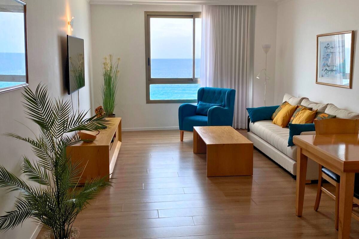 A modern living room with a wooden floor, a blue sofa, a wooden coffee table, and a TV mounted on the wall.