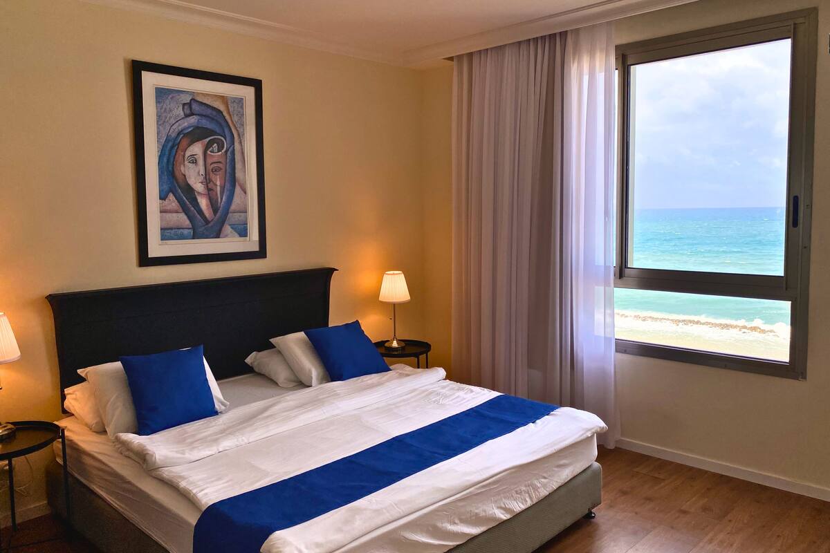 A hotel room with a bed, a framed artwork on the wall, a nightstand with a lamp, and a window with a beach view.