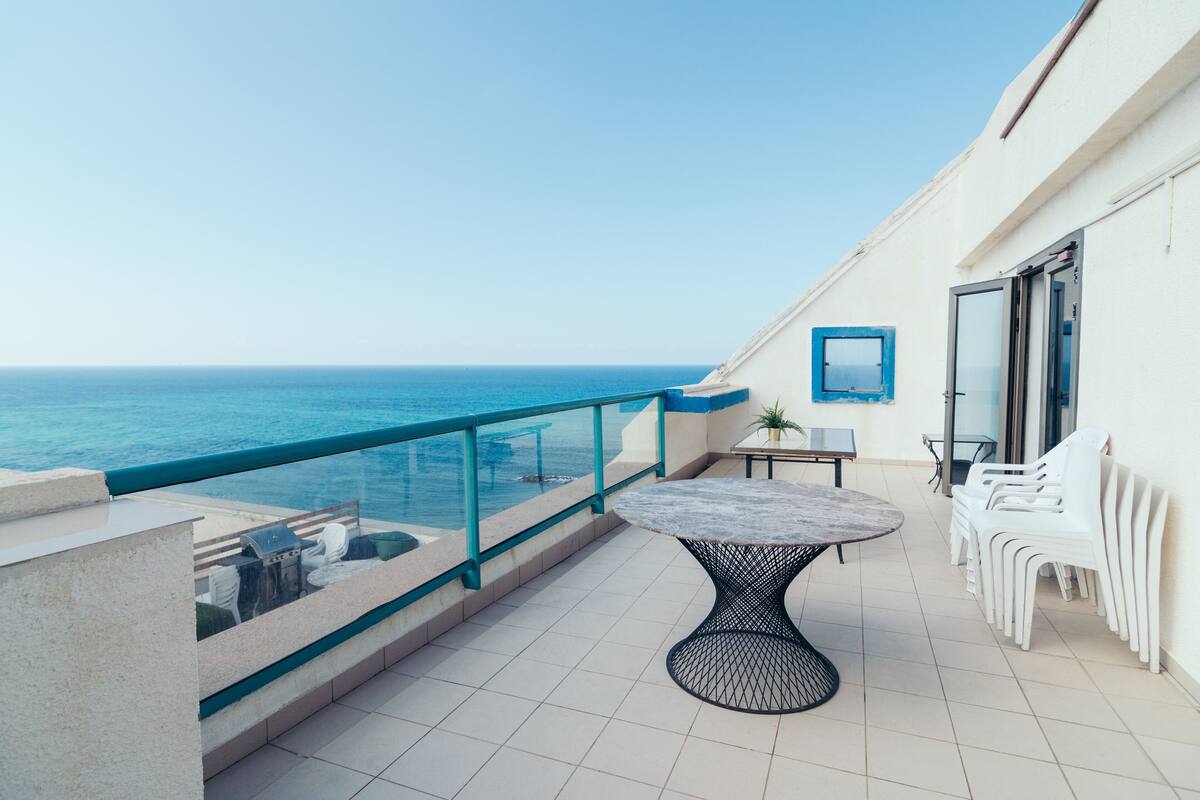 A balcony with a view of the sea, a round table, and white chairs.