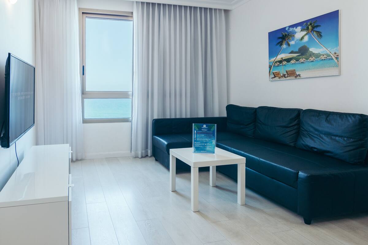 A modern living room with a dark green sofa, a white coffee table with a sign, a flat-screen TV, and a large window with curtains.