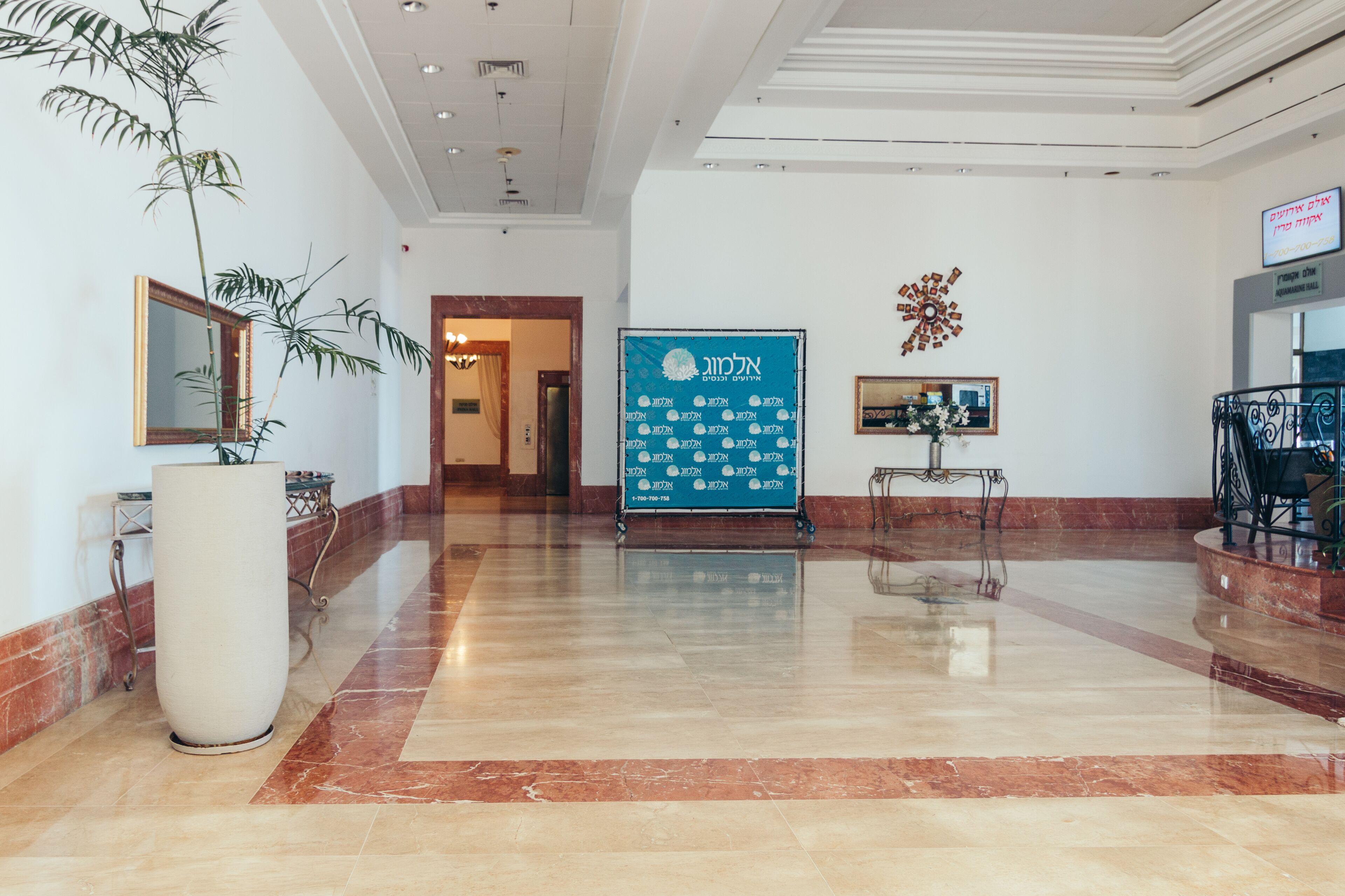 A spacious lobby with a polished marble floor, a large potted plant, a reception desk, and a blue sign with Hebrew text.