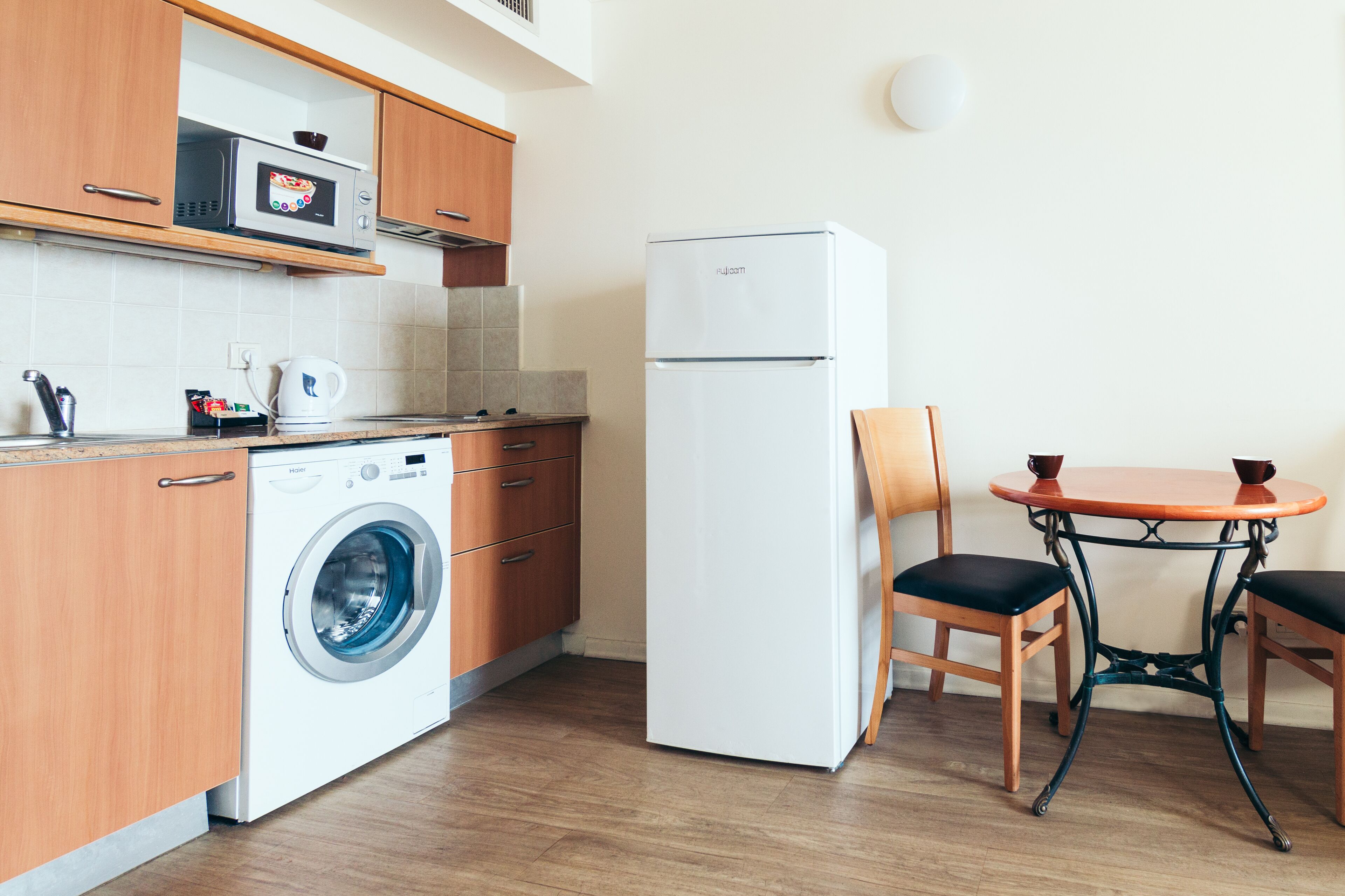 A compact kitchen with wooden cabinets, a washing machine, a refrigerator, and a small dining table with chairs.