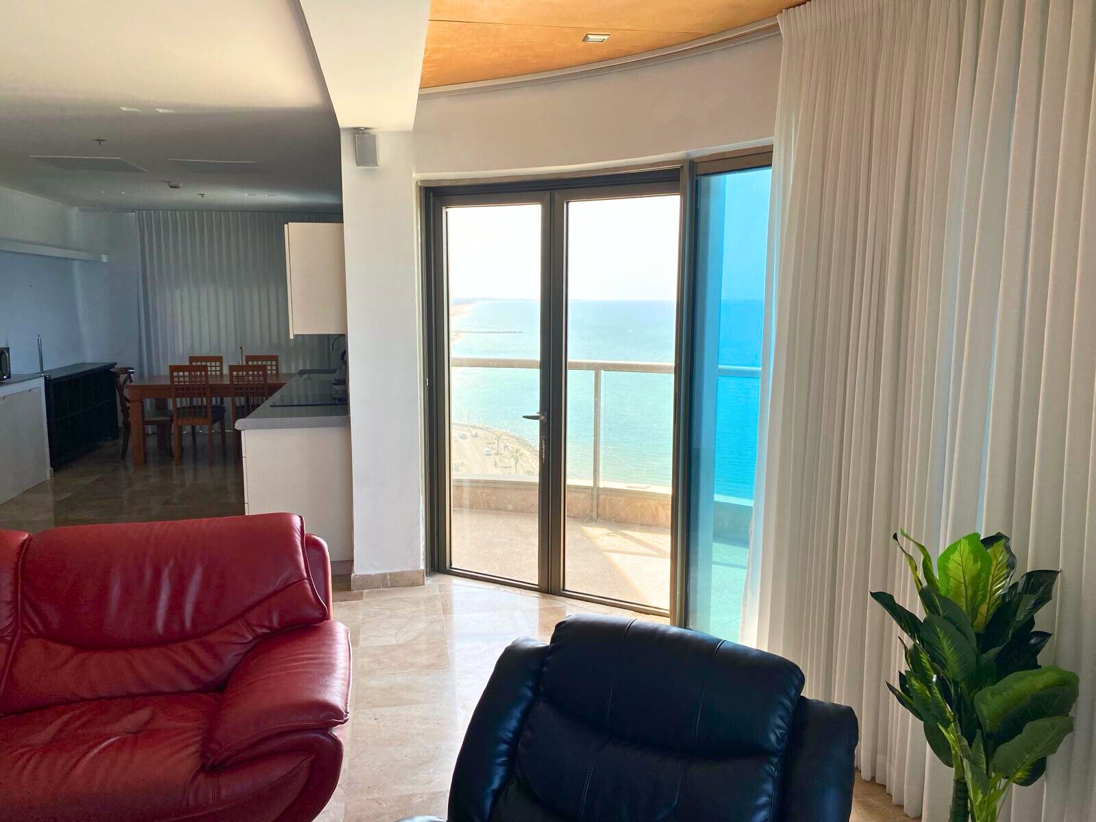 A modern living room with a red and blue leather sofa, a dining area with a table and chairs, and a large sliding glass door leading to a balcony with a view of the sea.