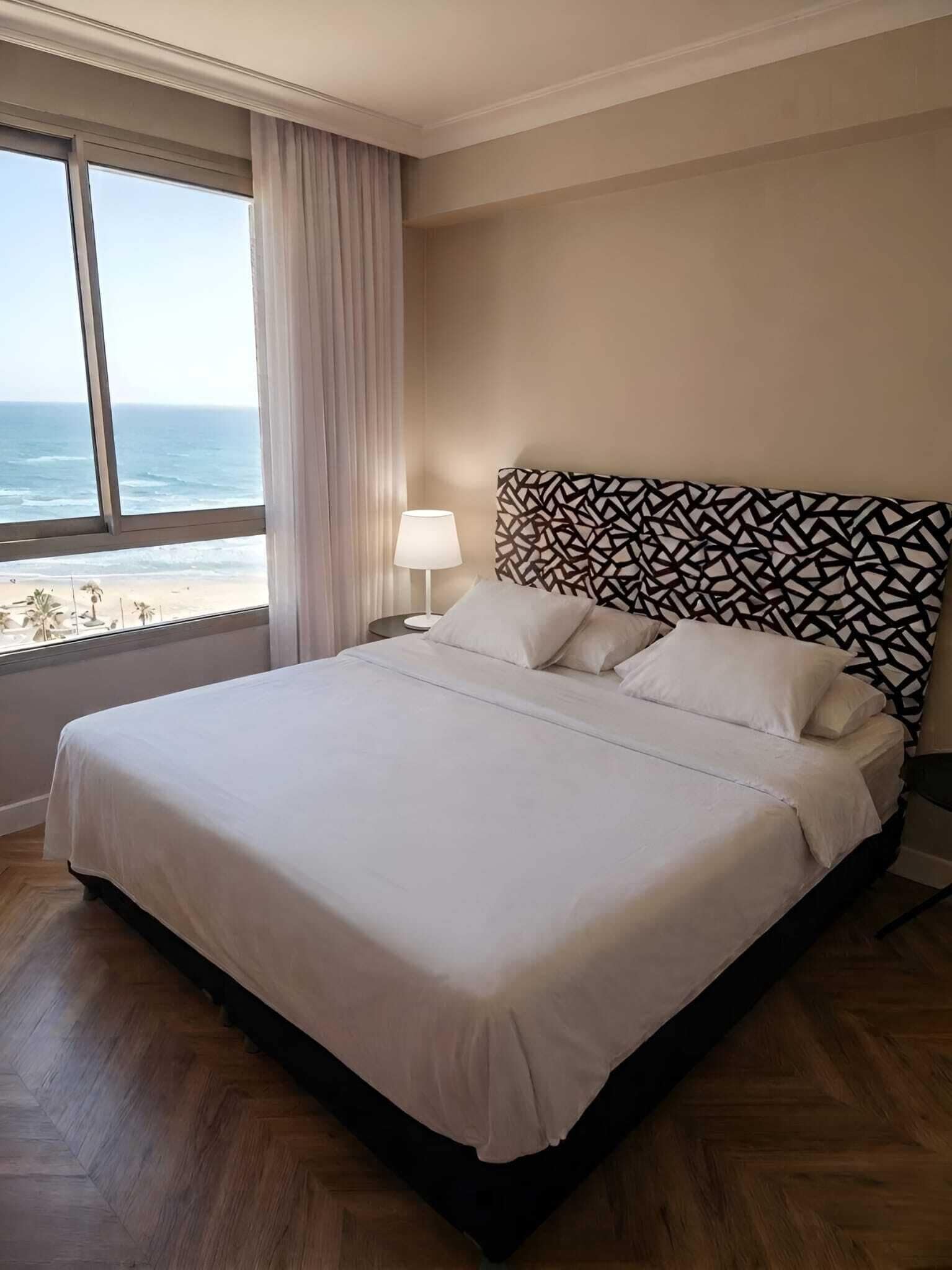 A hotel room with a large bed, a nightstand, a lamp, and a view of the ocean.