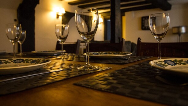 Une table à manger avec quatre verres à vin et des assiettes, placée dans une pièce à la lumière tamisée, avec des poutres apparentes et un luminaire fixé au mur.