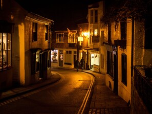 A narrow street at night with buildings on either side, illuminated by street lamps and shop lights. - Artists Cottage (Rye)