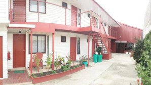 A two-story building with a red exterior, a covered front porch, and a small garden area with potted plants. - Hostal Maipu Street (Punta Arenas)