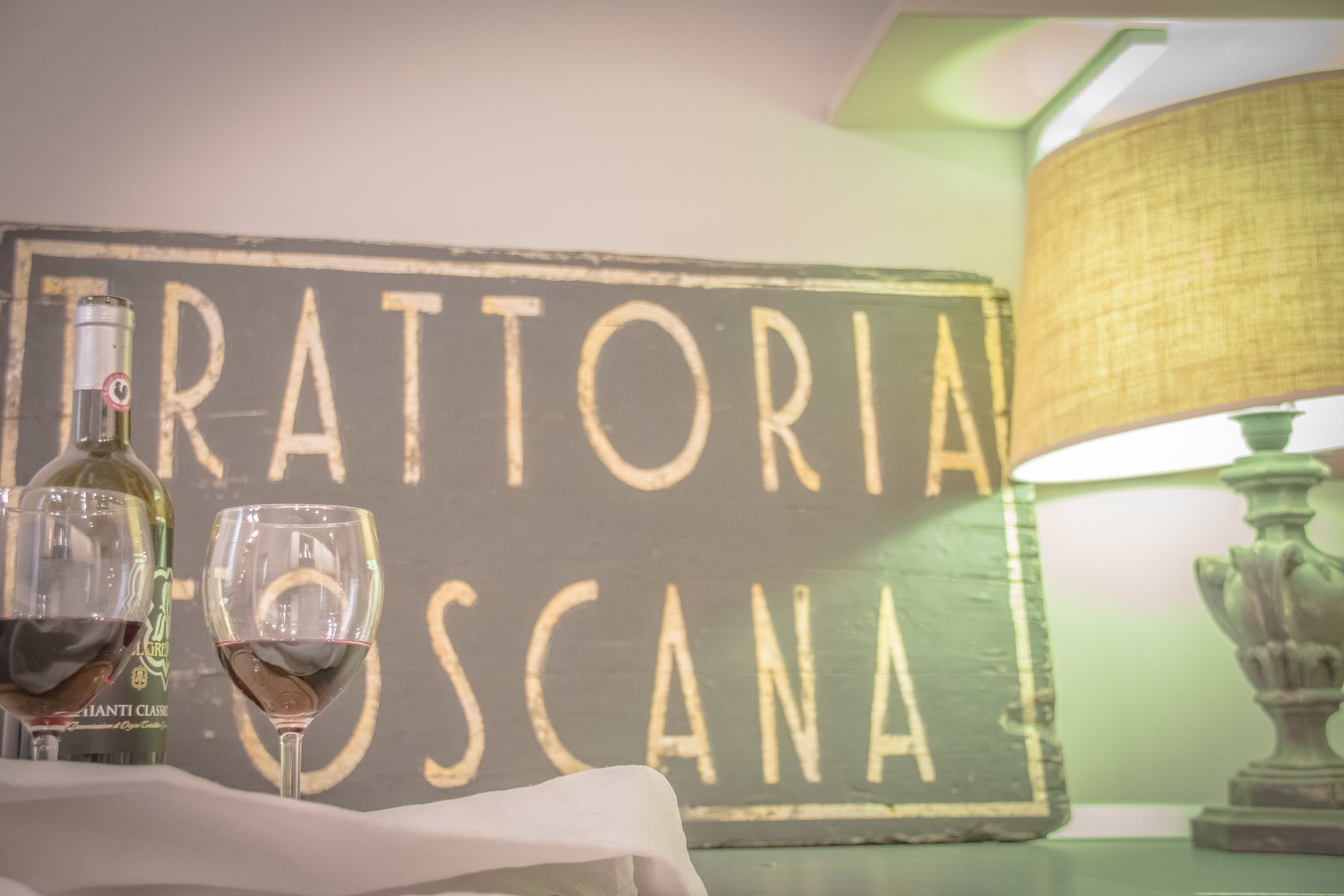 A table with two wine glasses and a bottle, a sign reading "TRATTORIA ROSCANA", and a lamp.