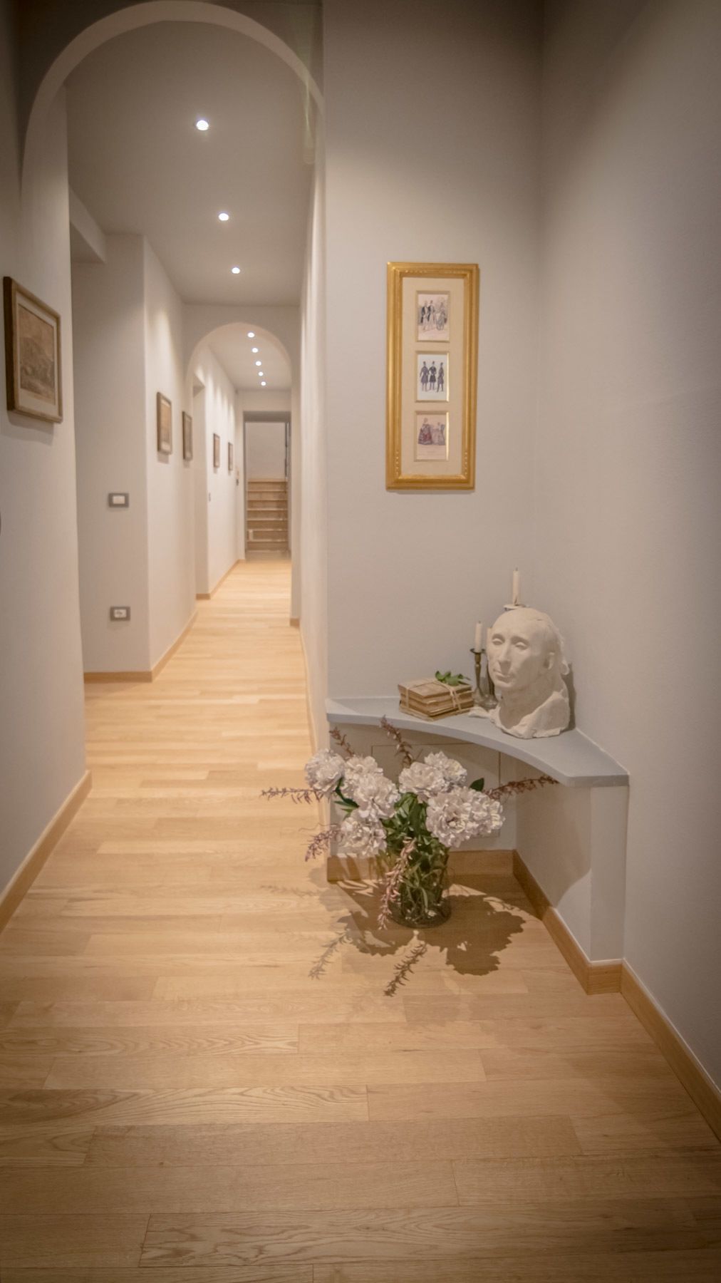 A hallway with wooden flooring, framed artwork on the walls, and a vase with flowers on a shelf.