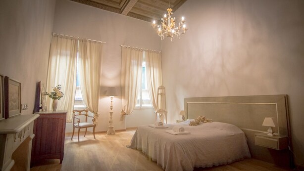 A bedroom with a large bed, a chandelier, a desk with a chair, and two windows with curtains. - Breathtaking 4BD Apt 5min Walk to Duomo (Florence)