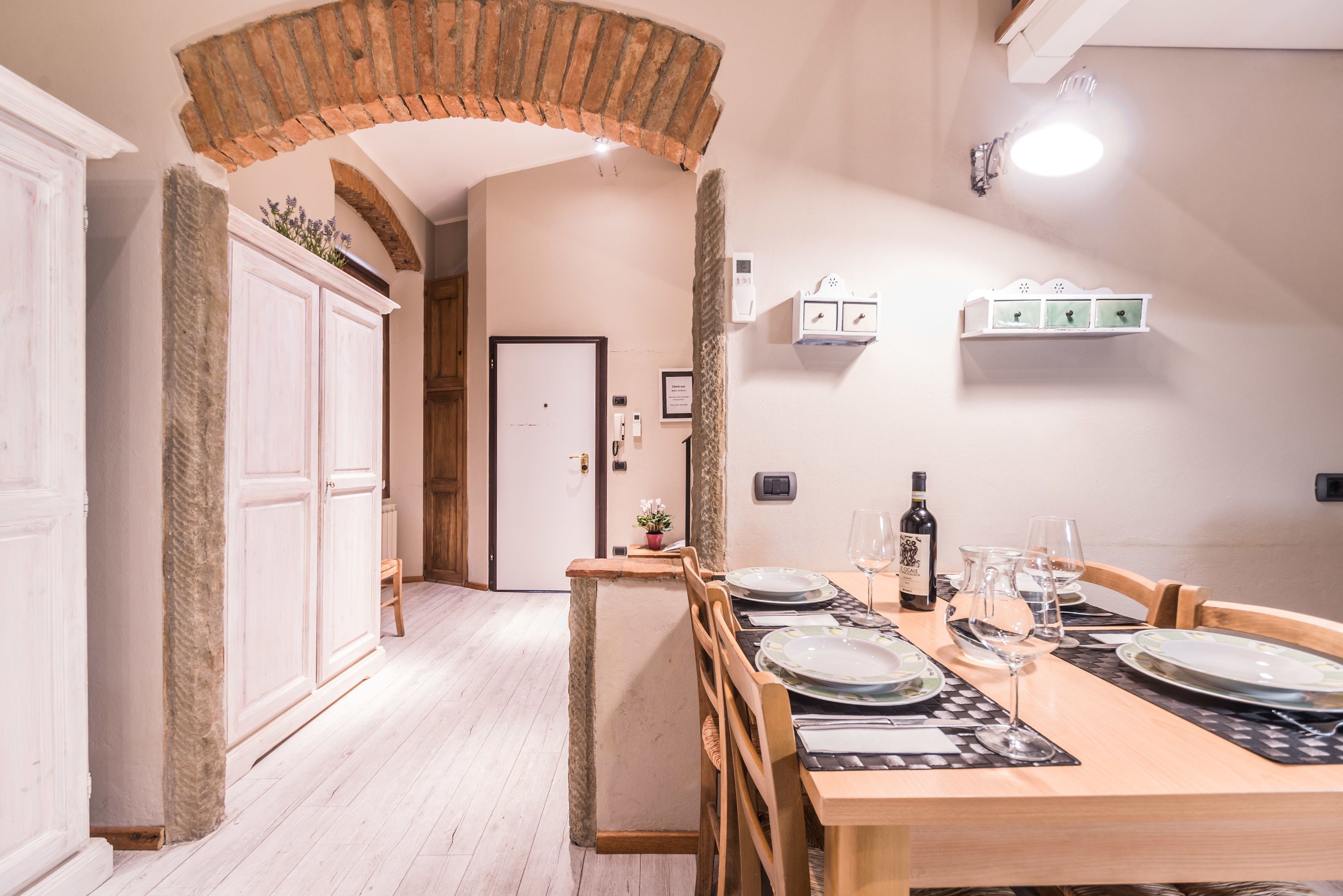 A dining area with a wooden table set for four, a brick archway, and a white cabinet with a plant.