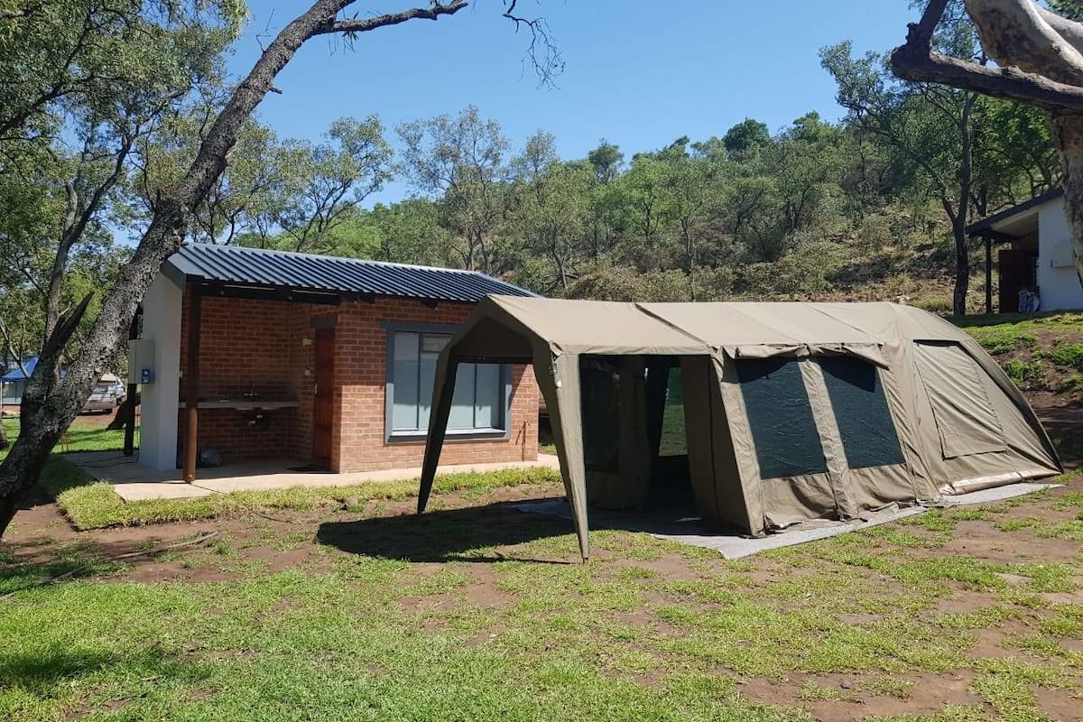 Un petit bâtiment en briques surmonté d’un toit équipé de panneaux solaires, une structure ressemblant à une tente, et une zone herbeuse avec des arbres.
