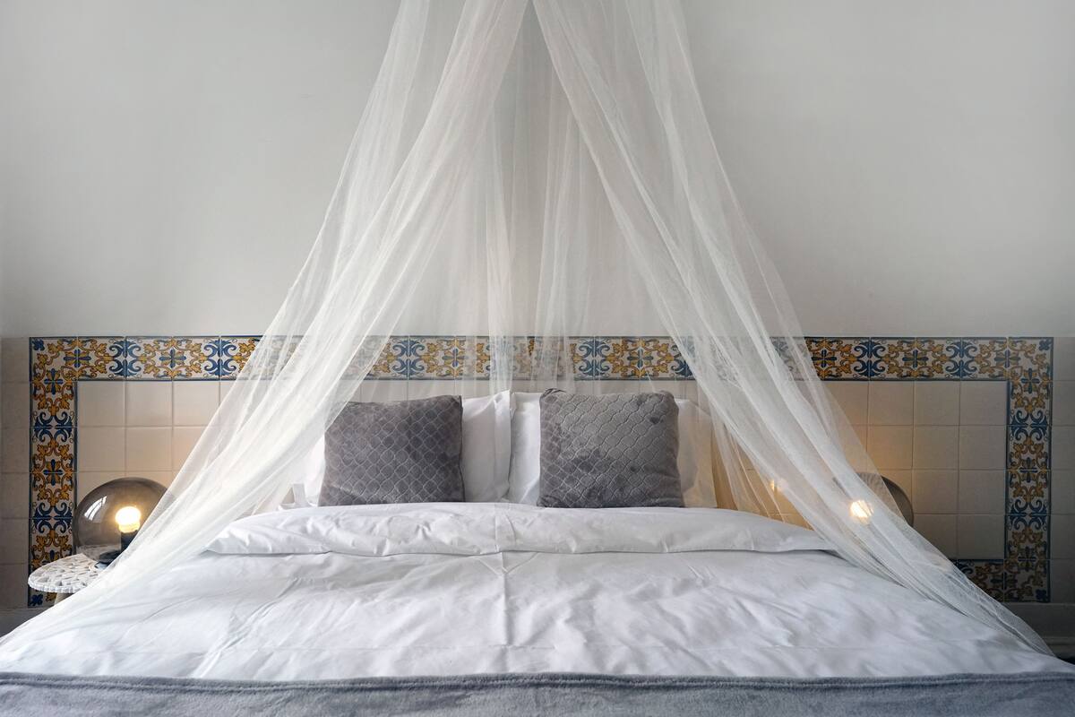 A bed with white linens and a canopy, a nightstand with a lamp, and a tiled wall with a decorative border.