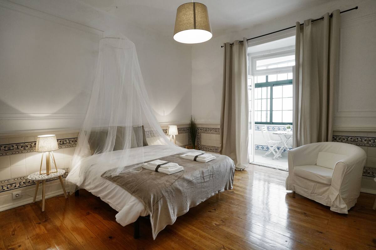 A bedroom with a bed, a chair, a bedside table, a lamp, and a window with curtains.