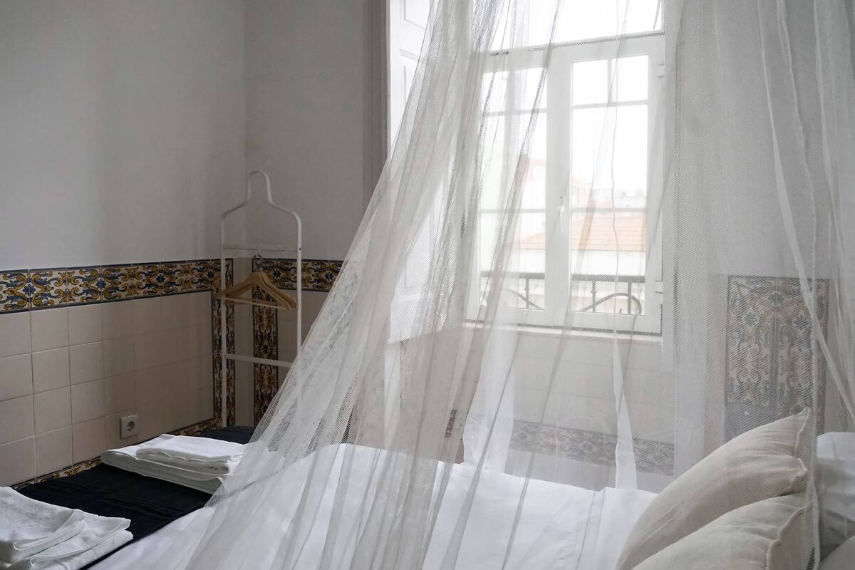 A bedroom with a bed, white curtains, a hanging light fixture, and a window with a view.