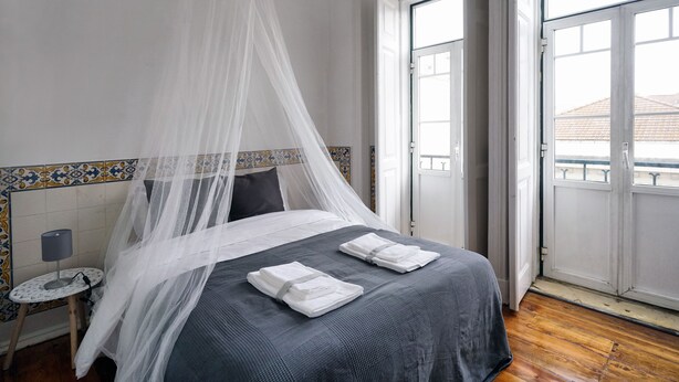 A neatly made bed with a grey bedspread and white towels, a bedside table with a lamp, and a window with a view of buildings. - 1924 (Lisbon)