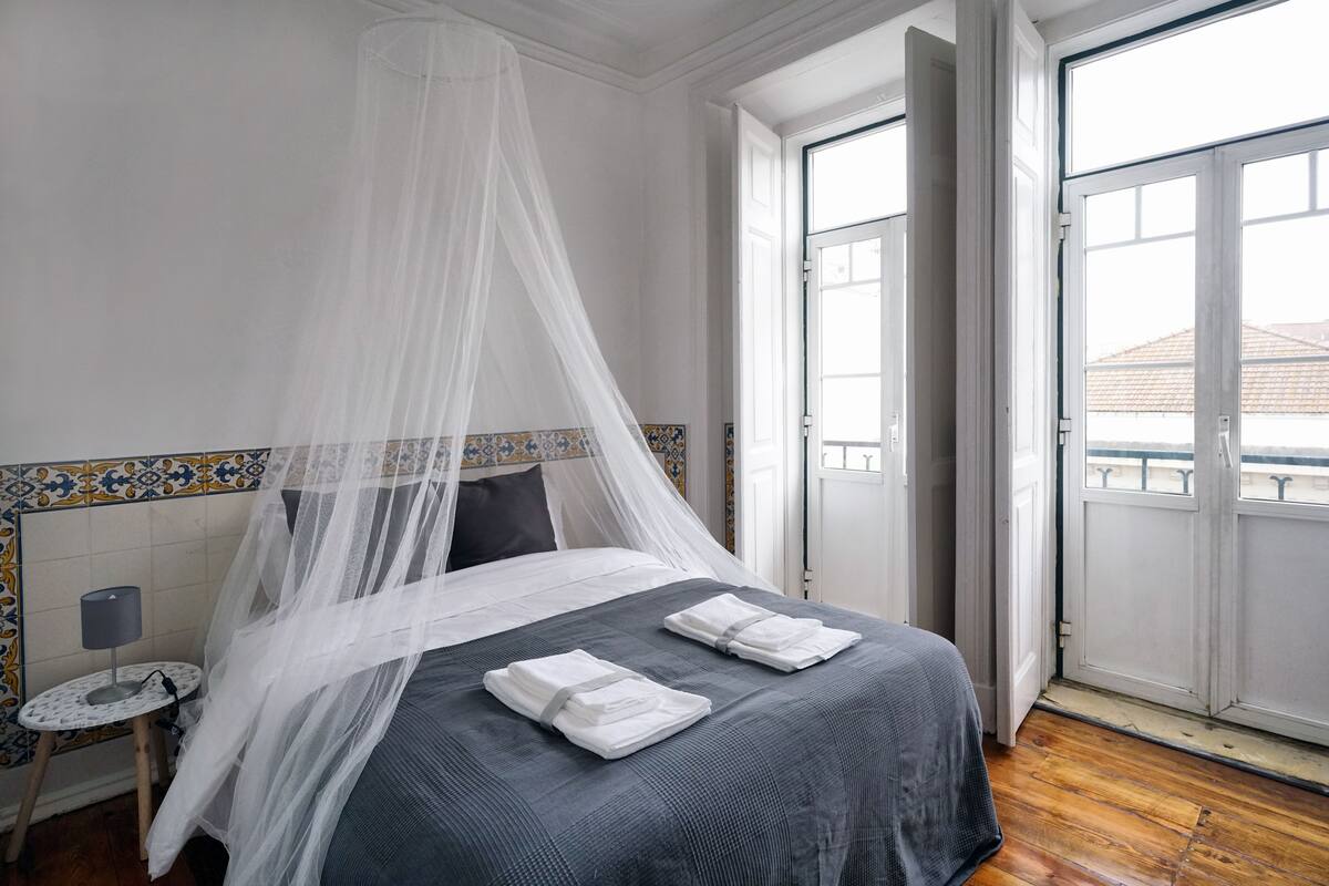 A neatly made bed with a grey bedspread and white towels, a bedside table with a lamp, and a window with a view of buildings.