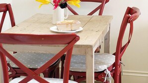Une table à manger ornée d’un vase de fleurs, une assiette avec une part de gâteau et des chaises rouges avec des coussins blancs.
