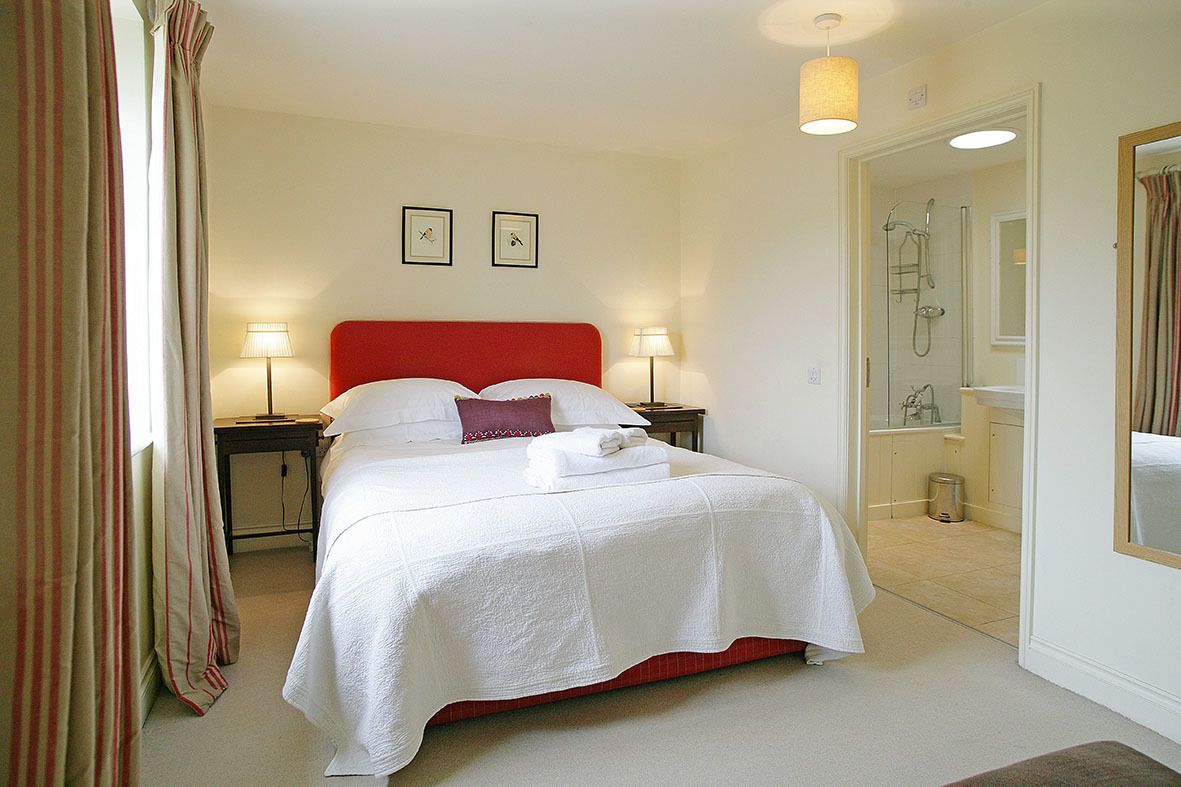 Une chambre d’hôtel avec une tête de lit rouge, du linge de lit blanc, deux lampes de chevet et un espace douche visible à travers une porte ouverte.