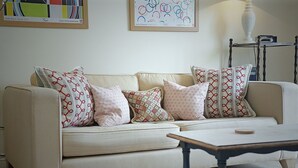 Un salon avec un canapé beige, des coussins à motifs, une table basse en bois et un tableau abstrait encadré accroché au mur.