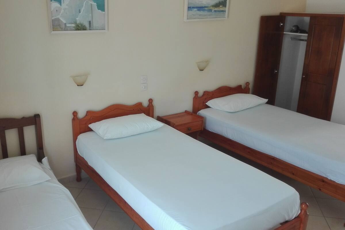A room with three wooden beds, each with white bedding. There are wall-mounted light fixtures and a wardrobe.