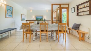 A dining area with a large table, wooden chairs, and a bench. There are wall-mounted lights, framed pictures, and a shelf with decorative items. - Beachy Barn (Eastbourne)