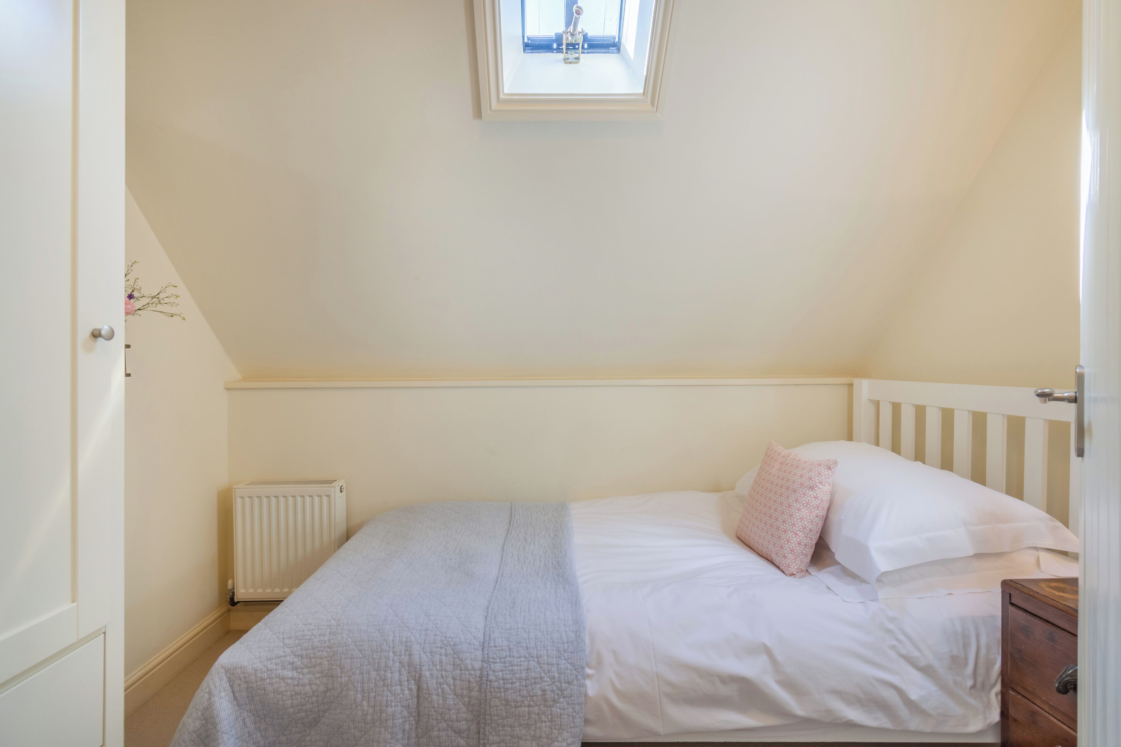Un dormitorio con una cama individual, un radiador blanco, un ventanal pequeño y una mesita de noche de madera.