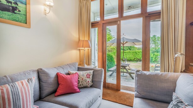 A living room with a grey sofa, a red pillow, and a lamp. There is a window with curtains and a view of a garden. - Beachy Barn (Eastbourne)
