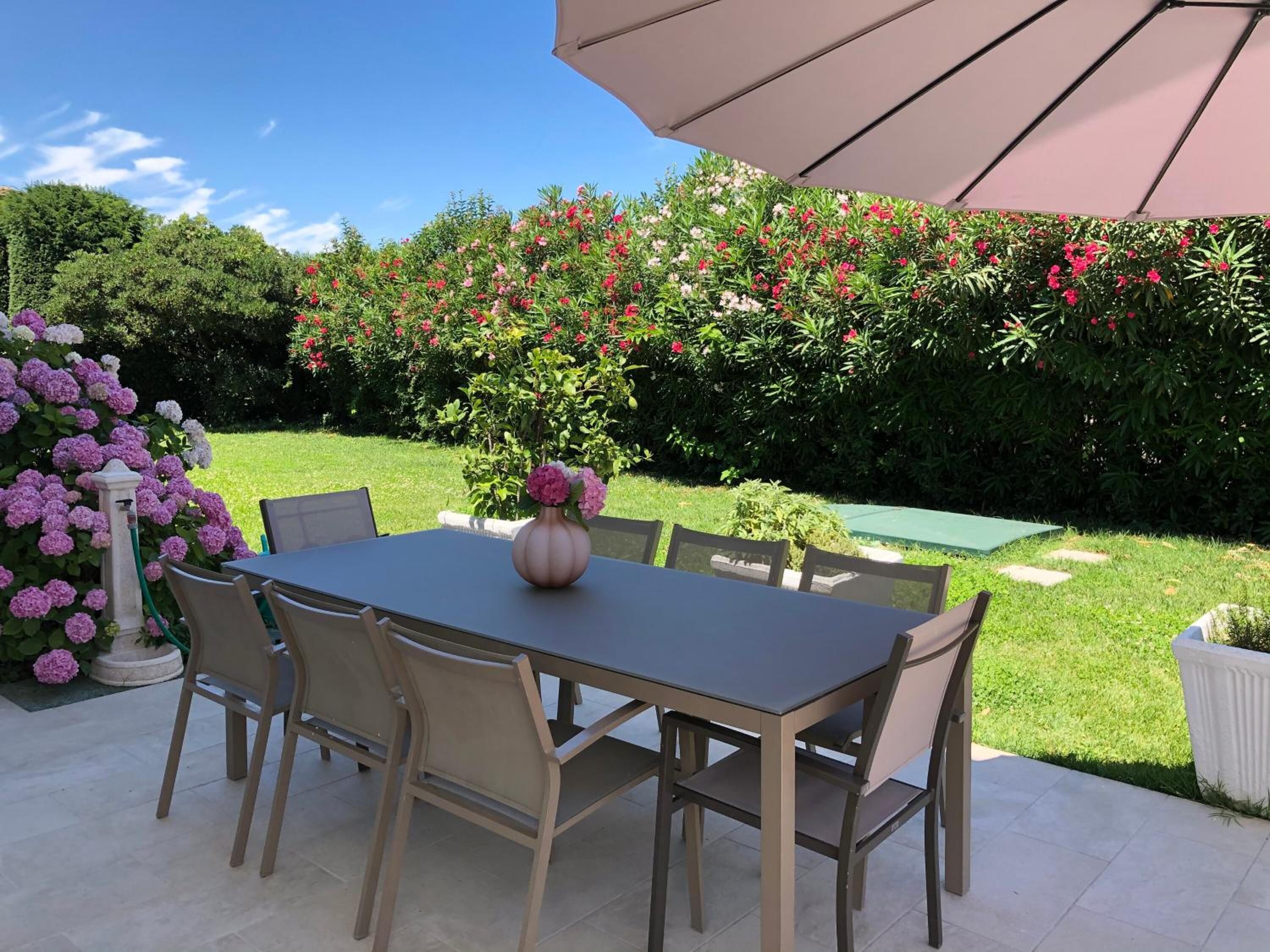 En udendørs terrasse med et bord og stole, omgivet af frodigt grønt og blomstrende planter.