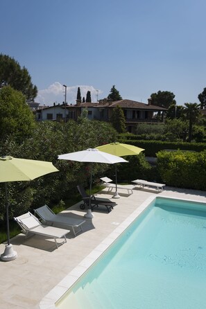 A pool area with lounge chairs and umbrellas, surrounded by greenery and a building in the background. - Bed & Breakfast da Beatrice (Sirmione)