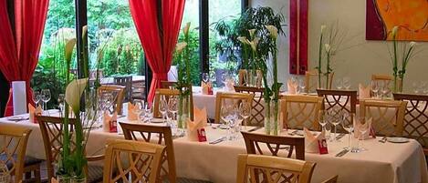 A dining area with tables set for multiple guests, featuring chairs with a lattice back design, and large windows with red curtains.