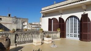 A courtyard with a stone balustrade, potted plants, and a building with arched windows and brown shutters. - B&B Casa Resci (Tricase)