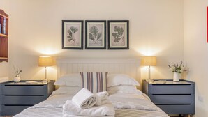 Een net opgemaakt bed met witte lakens, twee nachtkastjes met lampen, en drie ingelijste botanische prints aan de muur.