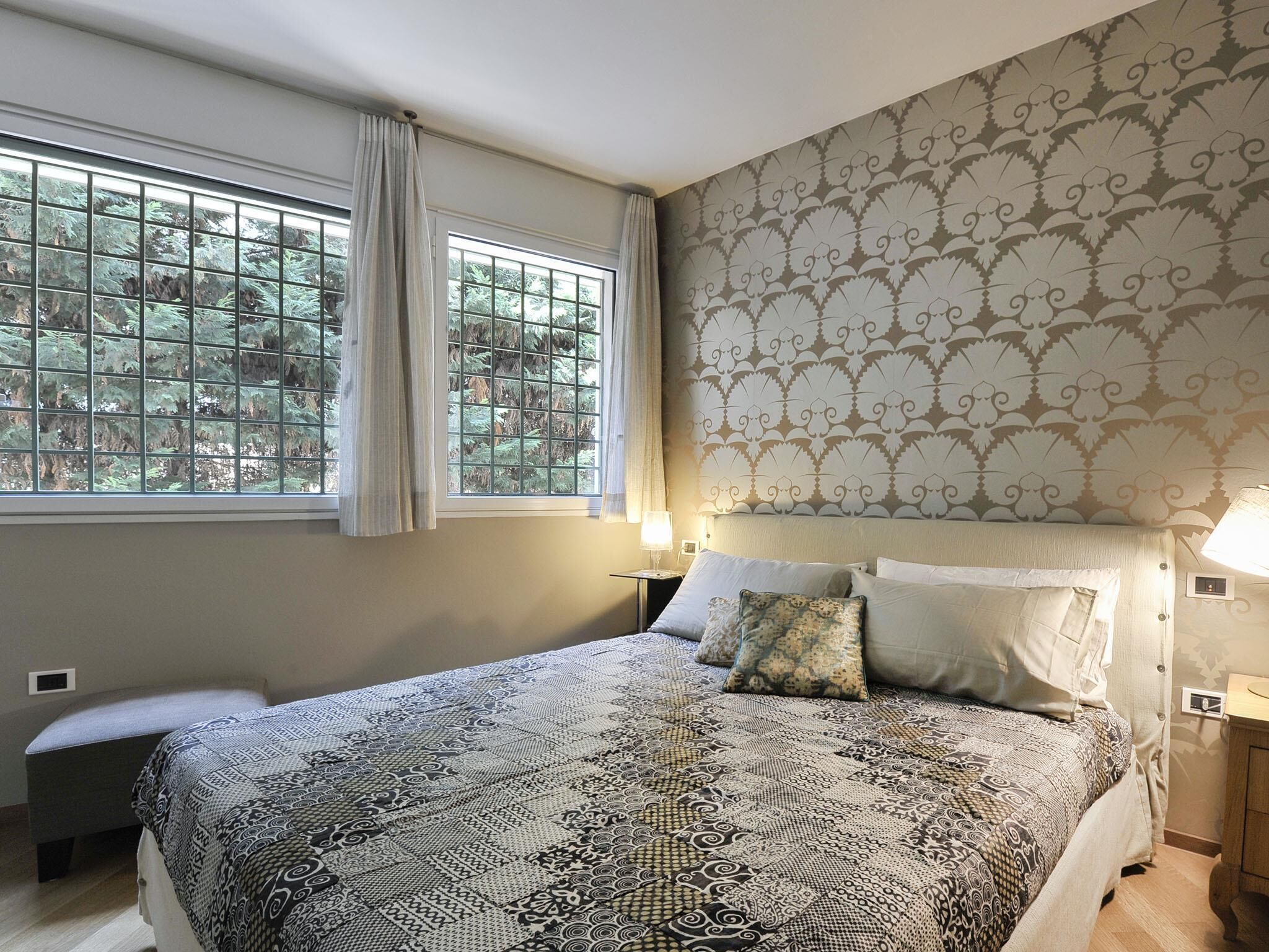 A bedroom with a large bed, a window with curtains, a patterned wall, and a bedside table with a lamp.
