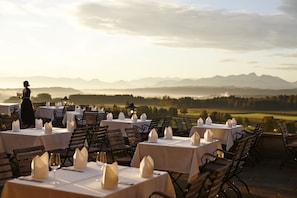 Un área de comedor al aire libre con mesas preparadas para una comida, con vistas a un paisaje pintoresco con montañas y campos.