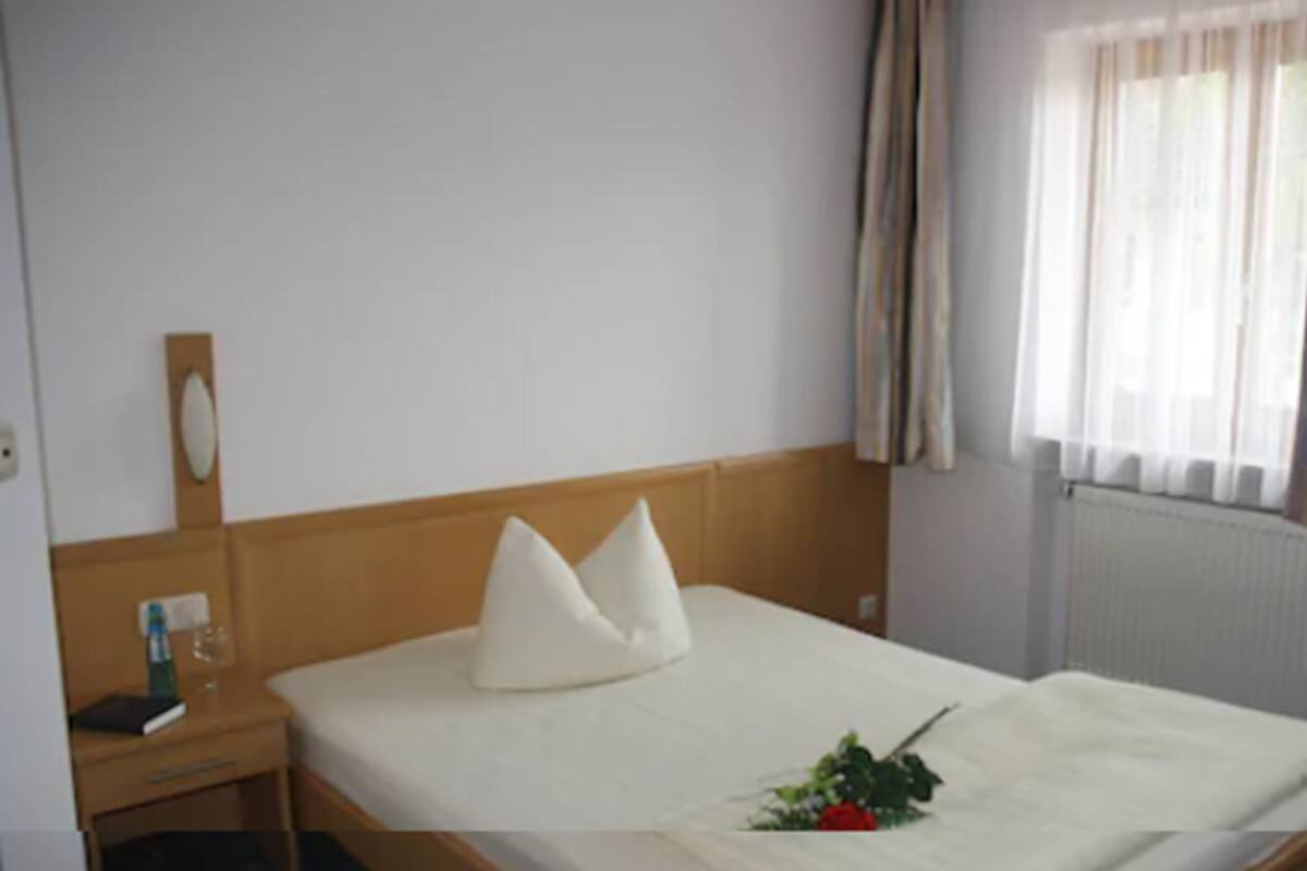 A hotel room with a wooden headboard, a single bed with white bedding, a bedside table with a lamp, and a window with sheer curtains.
