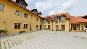 A yellow building with a red roof, multiple windows, and a paved courtyard. - Hotel Garni Jagermo (Grasbrunn)