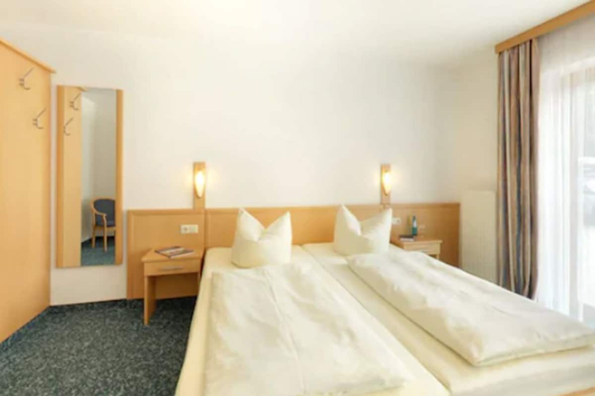 A hotel room with a large bed, two bedside tables, a chair, a mirror, and a window with curtains.