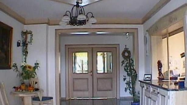 A spacious living area with a high ceiling, a chandelier, a dining table with chairs, and a kitchen area with cabinets. - Hotel Garni Jagermo (Grasbrunn)