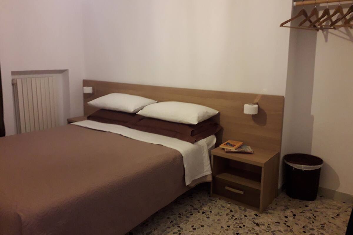 Un dormitorio con una cama, una mesita de noche, un cubo de basura y un estante de madera con un libro.