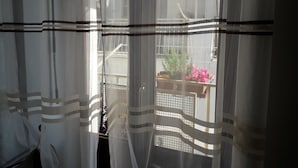 Ein Zimmer mit transparenten Vorhängen, Blick auf einen Balkon mit Topfpflanzen und Blumen, sowie ein Fenster mit Gittermuster.