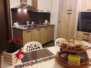 Una cocina con armarios de madera, una estufa y un lavamanos. Hay una mesa con mantel floral y una bandeja de pan junto a un recipiente amarillo.