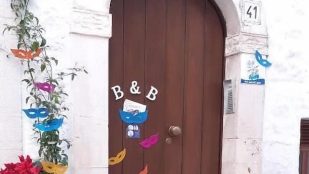 Eine braune Tür mit der Aufschrift "B&B" darüber, ein kleines Schild mit einer Maske und eine Blumenkomposition auf den Stufen.