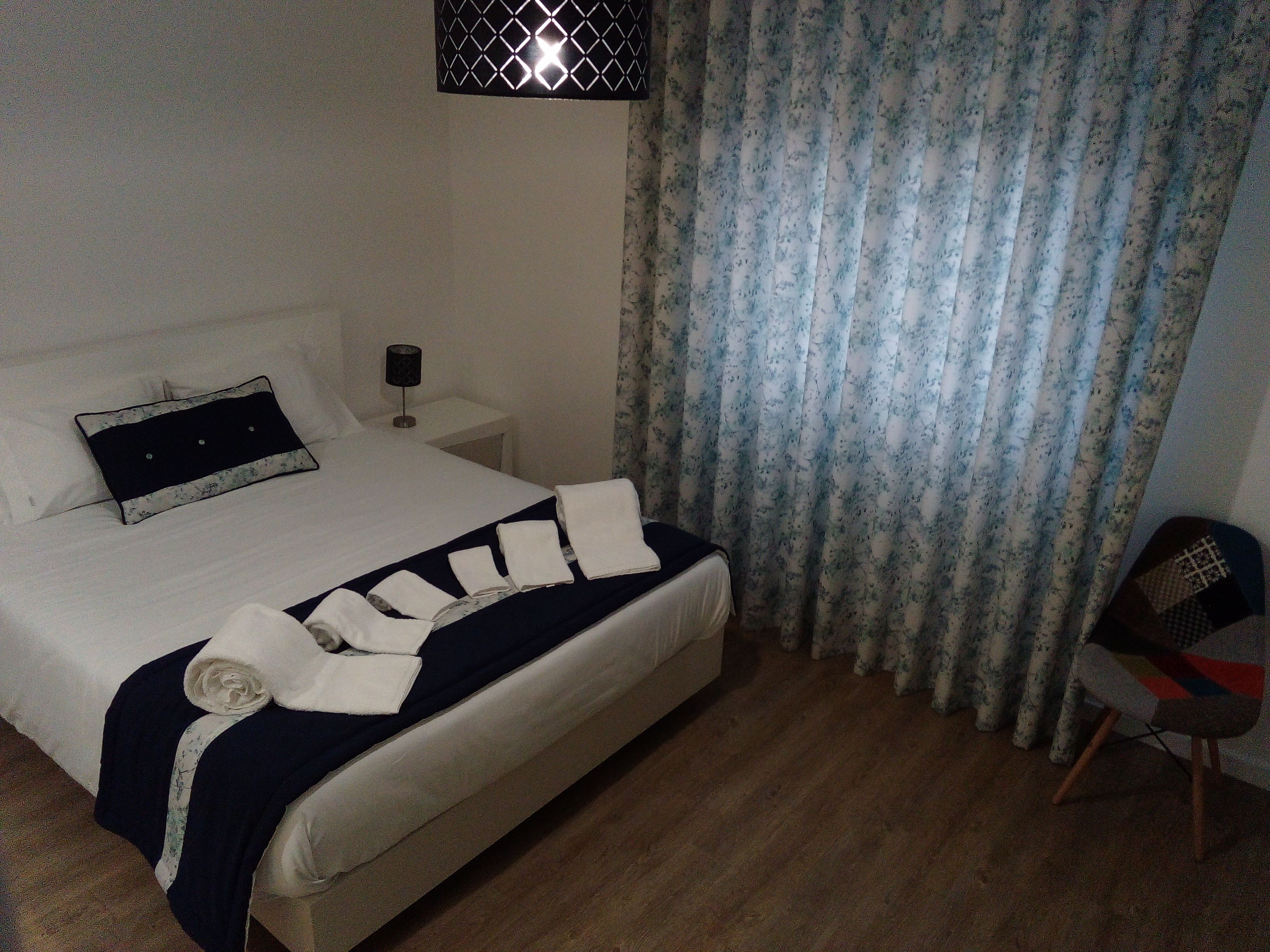 Uma cama bem arrumada com um cobre-leito azul escuro e toalhas brancas, uma mesa de cabeceira com abajur e uma cortina florida.