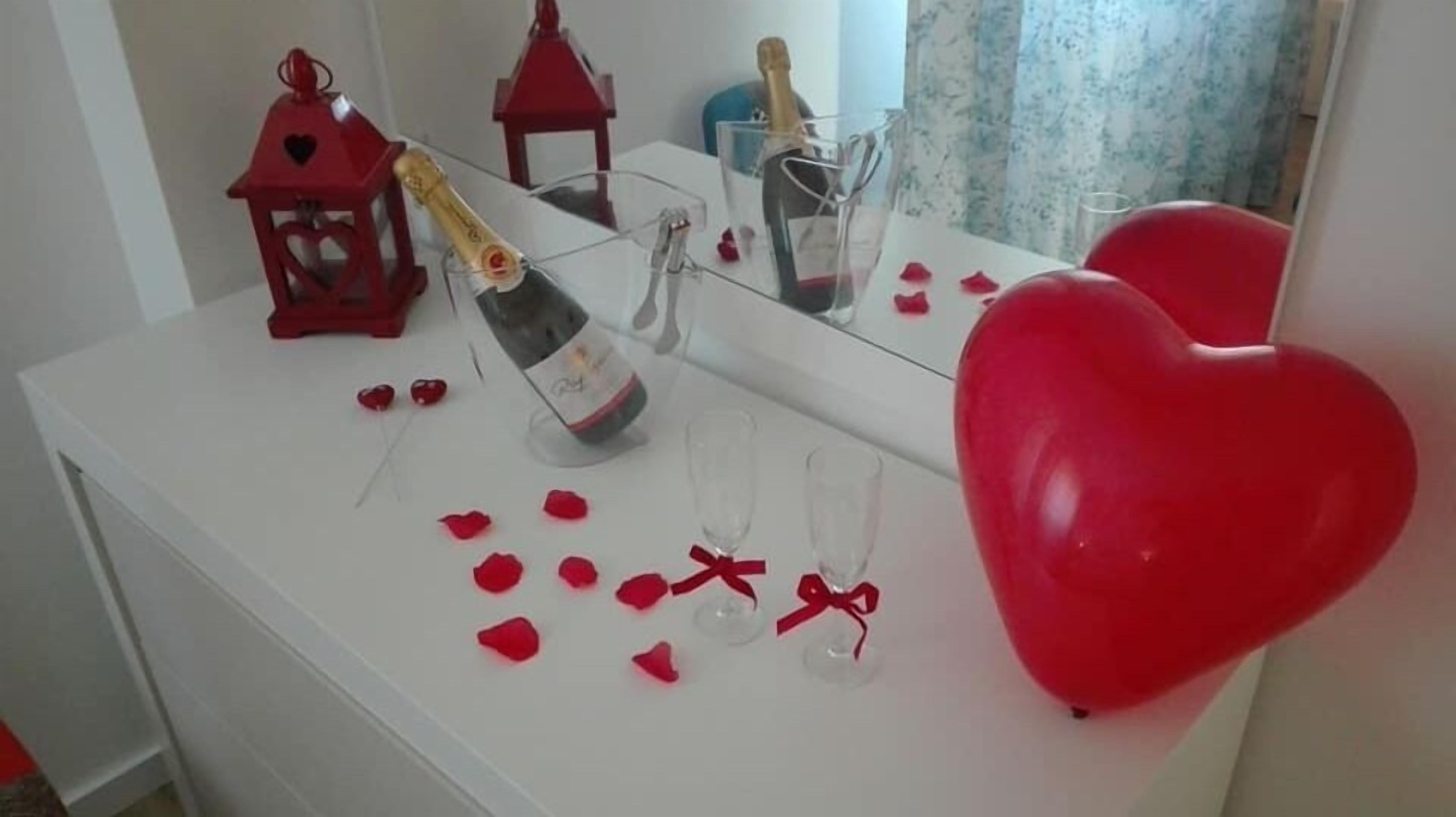 A white table with a bottle of champagne, two glasses, a red heart-shaped balloon, and scattered rose petals.