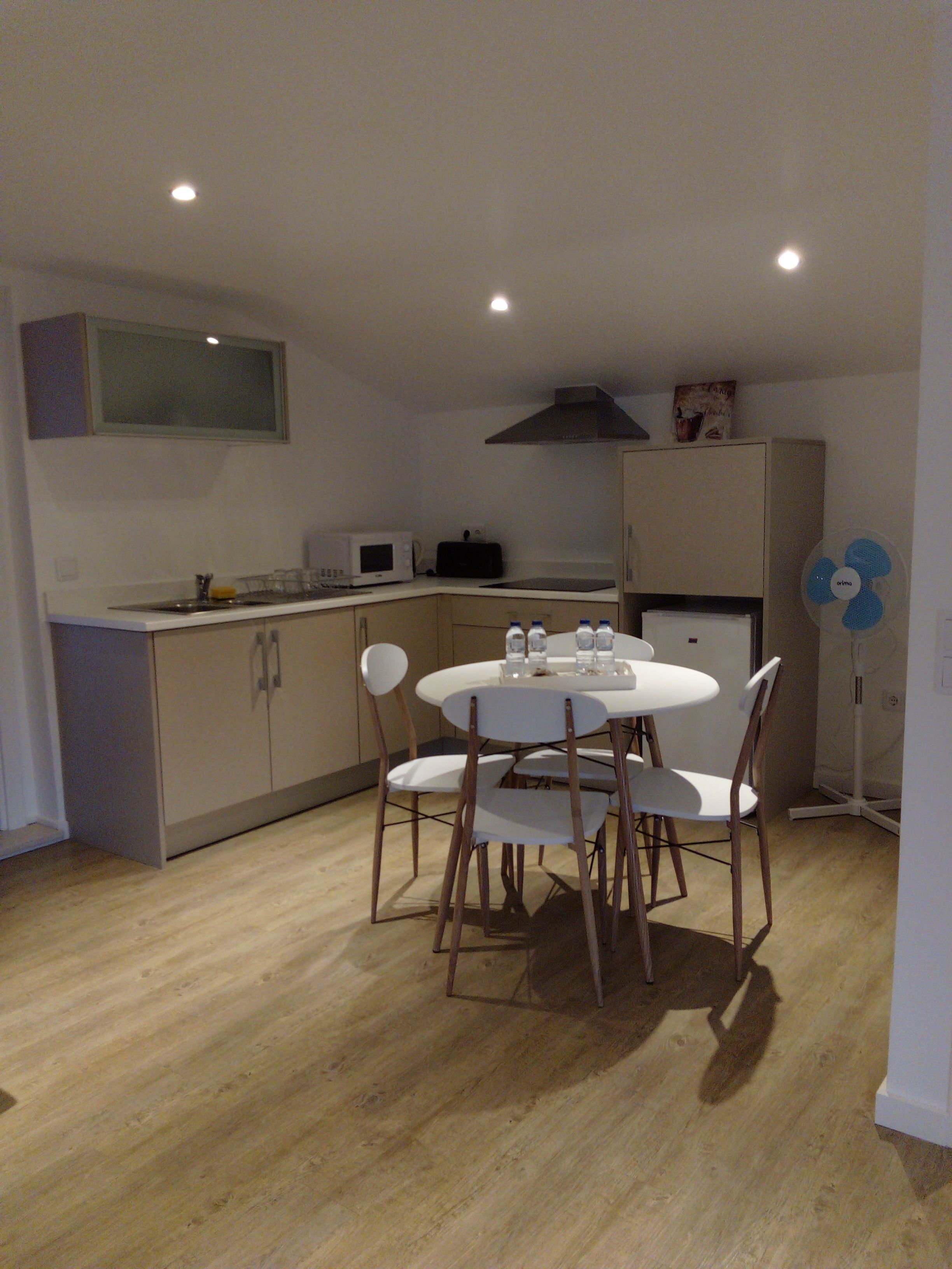 A compact kitchen with a dining table and chairs, a microwave, a small refrigerator, and a fan.