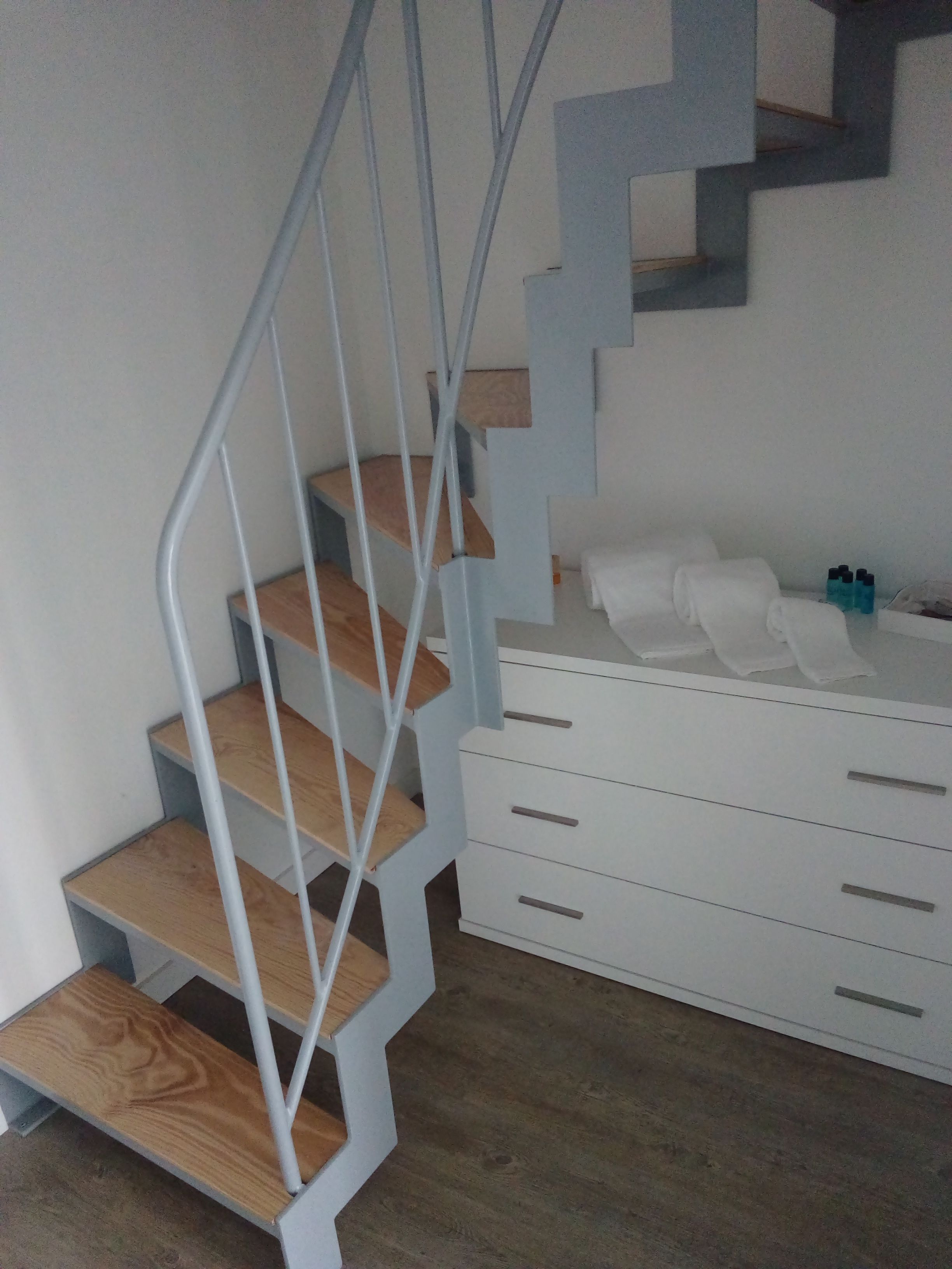 A modern staircase with wooden steps and a metal railing, leading to a white dresser with drawers and a small table with bottles.