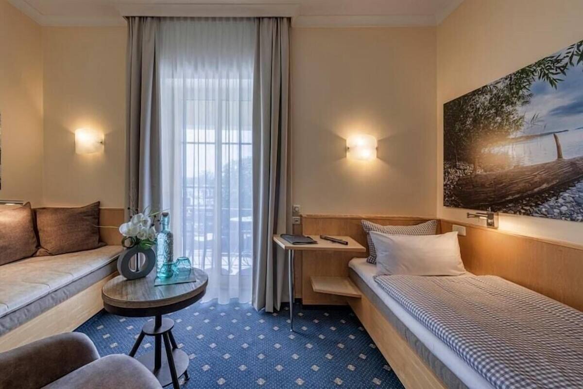 A hotel room with a sofa, a bed, a small table with a vase, a desk, and a painting on the wall.